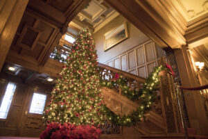 A tall Christmas tree lit with multi-color lights and ornaments stands in front of a wooden staircase and railing.