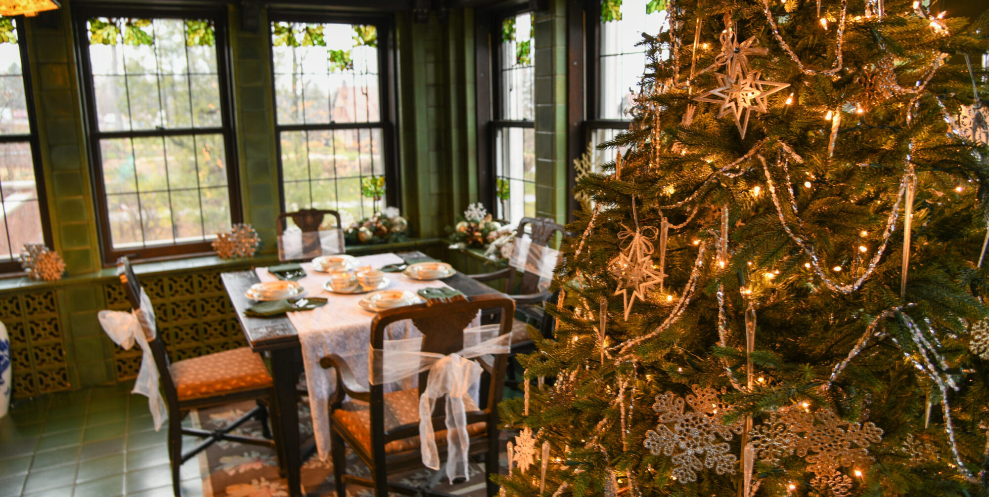 A Christmas tree decroated with lights, ornaments, and white snowflakes stands in front of a plated breakfast table and glass windows.