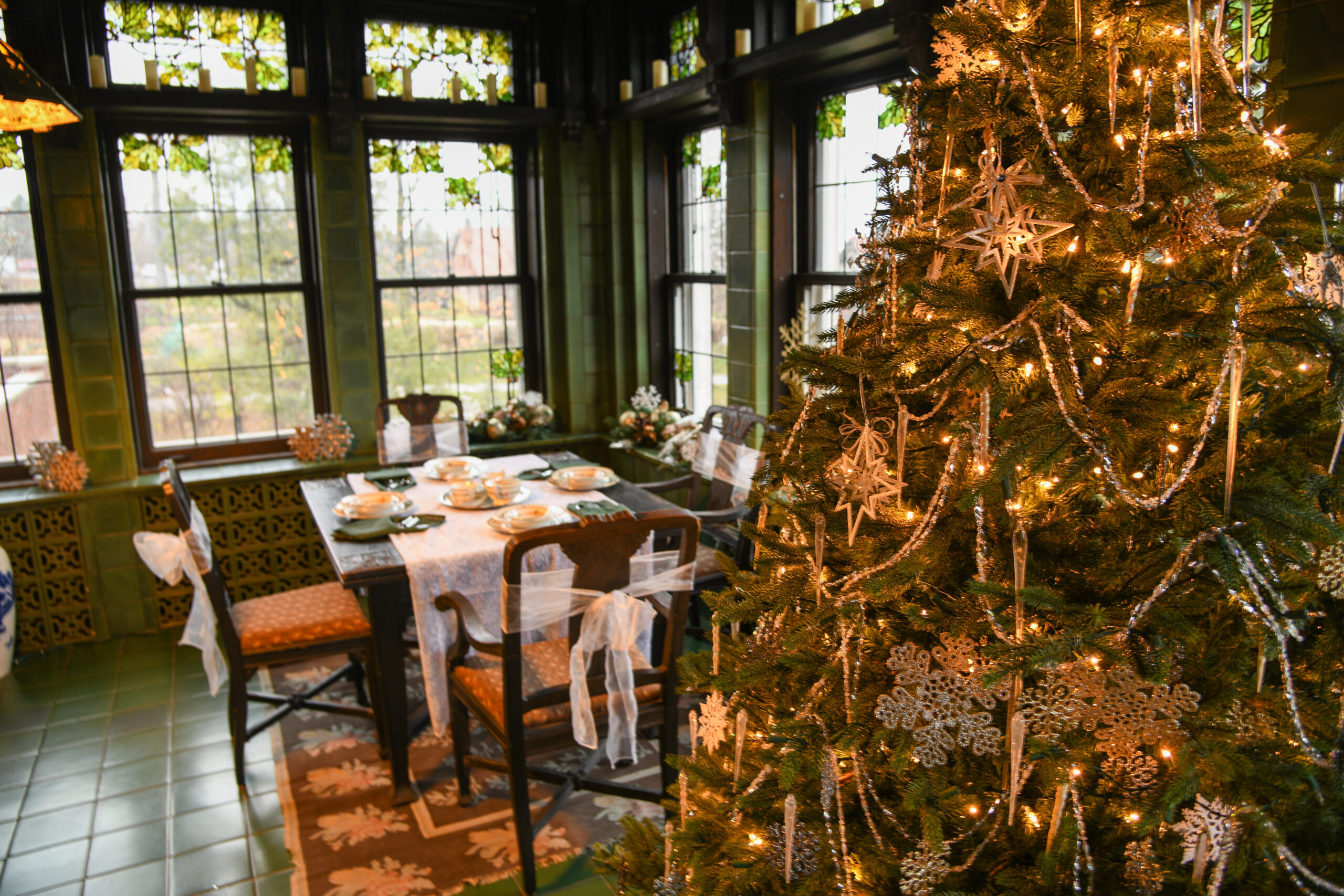 A Christmas tree decroated with lights, ornaments, and white snowflakes stands in front of a plated breakfast table and glass windows.