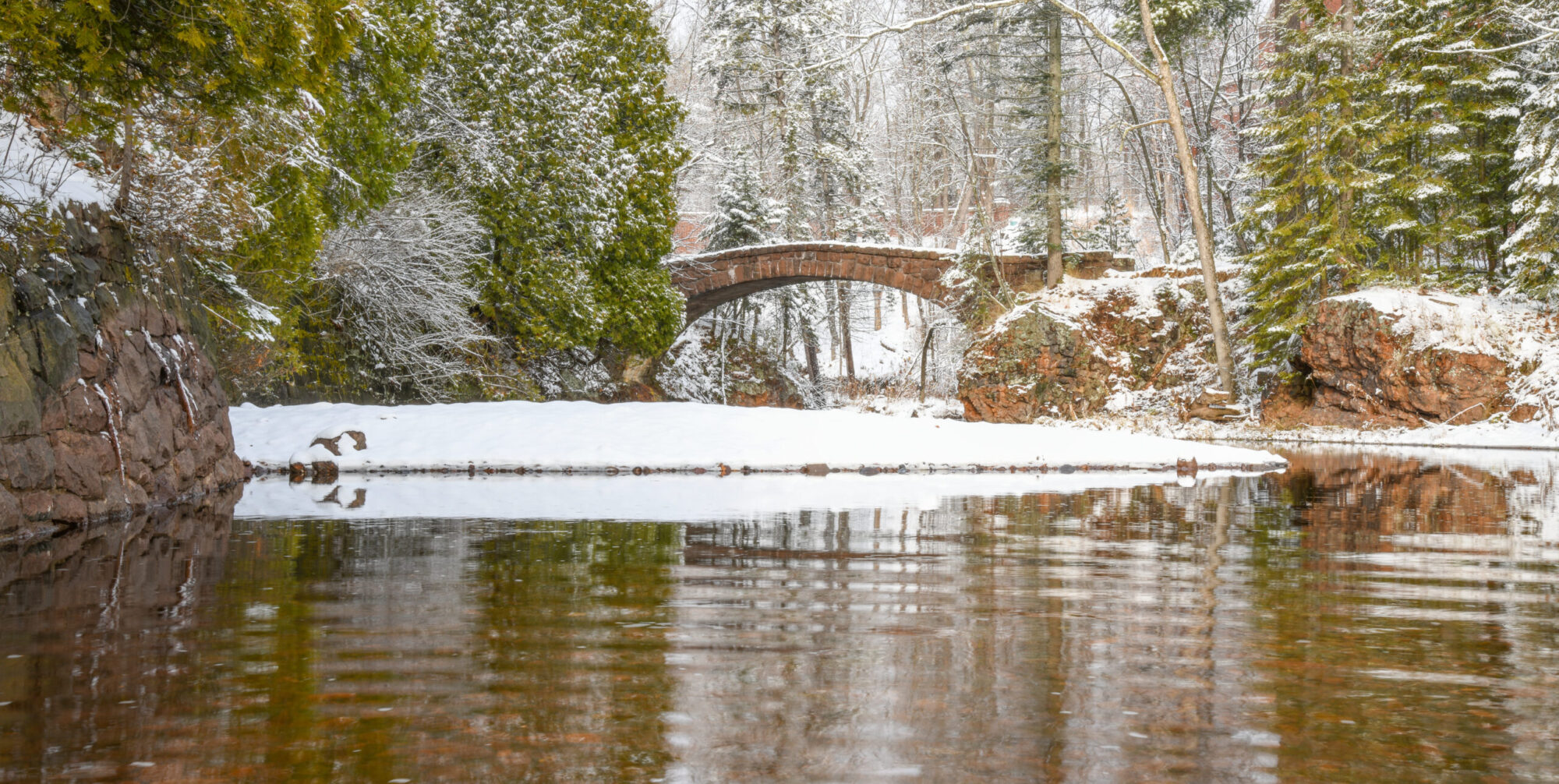 A bridge crosses over a river with snow on it and evergreen trees covered in snow on either side.