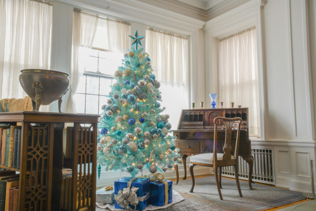 A blue Christmas tree stands decorated with ornaments and lights in front of a wooden shelf and with a wooden desk and chair in the background. The tree is in a brightly lit white room with windows behind it.