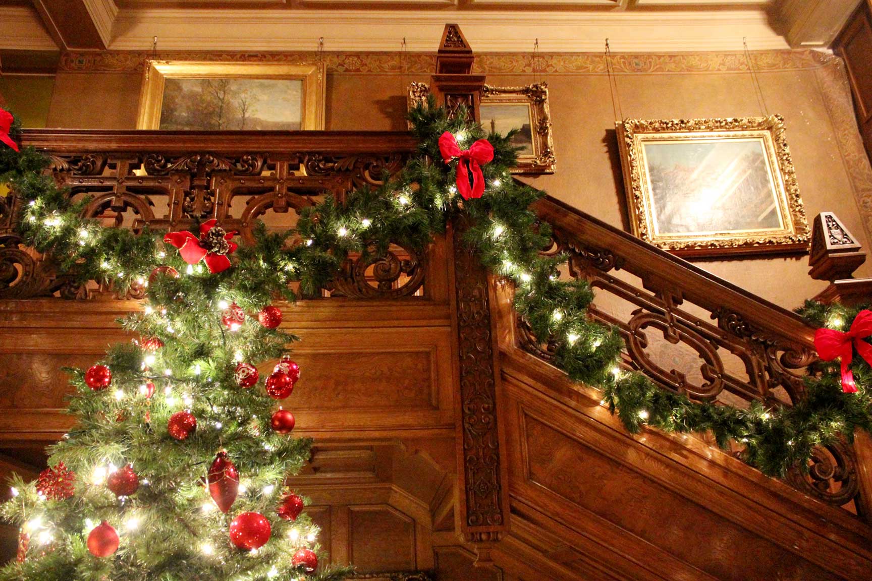 Green garland with white lights and red bowties lines wooden railings of a staircase with a large green tree in the foreground lit with white lights and red ornaments.