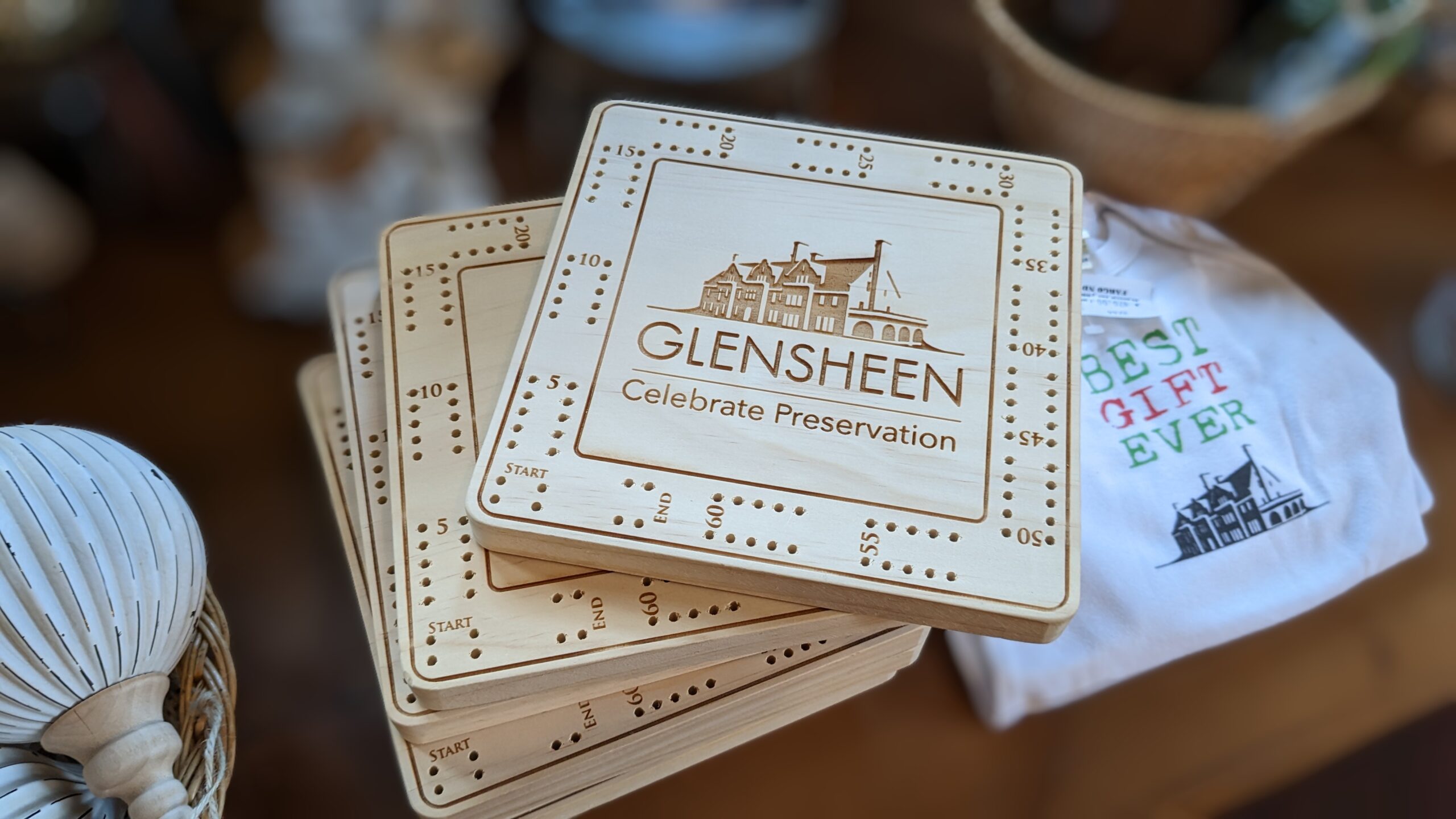 Wooden cribbage boards with the Glensheen logo sit on a table with a white t shirt in the background.