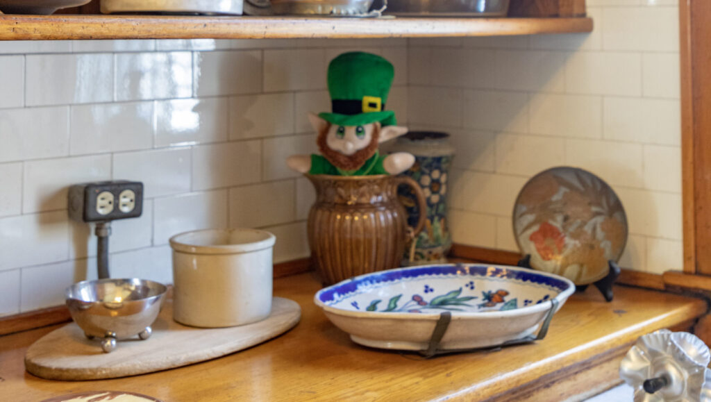 A green leprechaun sits in a vase on a wooden counter with dishes.