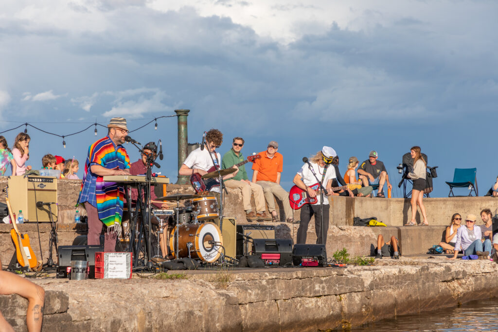 Members of the bad Damien perform music on a concrete pier in front of people on the water and on the pier.