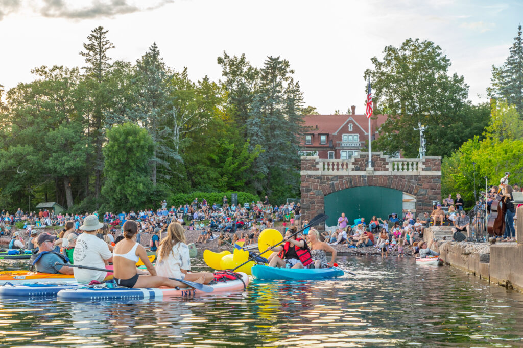 Kayakers and a person on a yellow rubber duck floatie are in the water in front of a brick boathouse and mansion with a pier on the right side of the image.