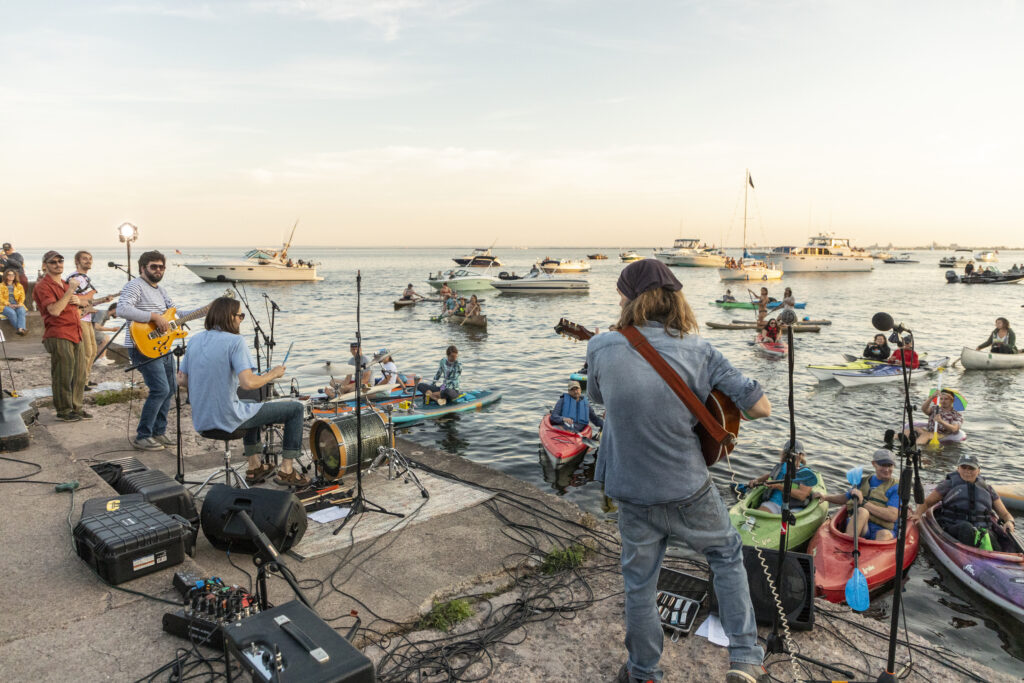 Teague Alexy and the Common Thread perform music on a concrete pier in front of kayakers and boaters in the water with the sun setting in the background.