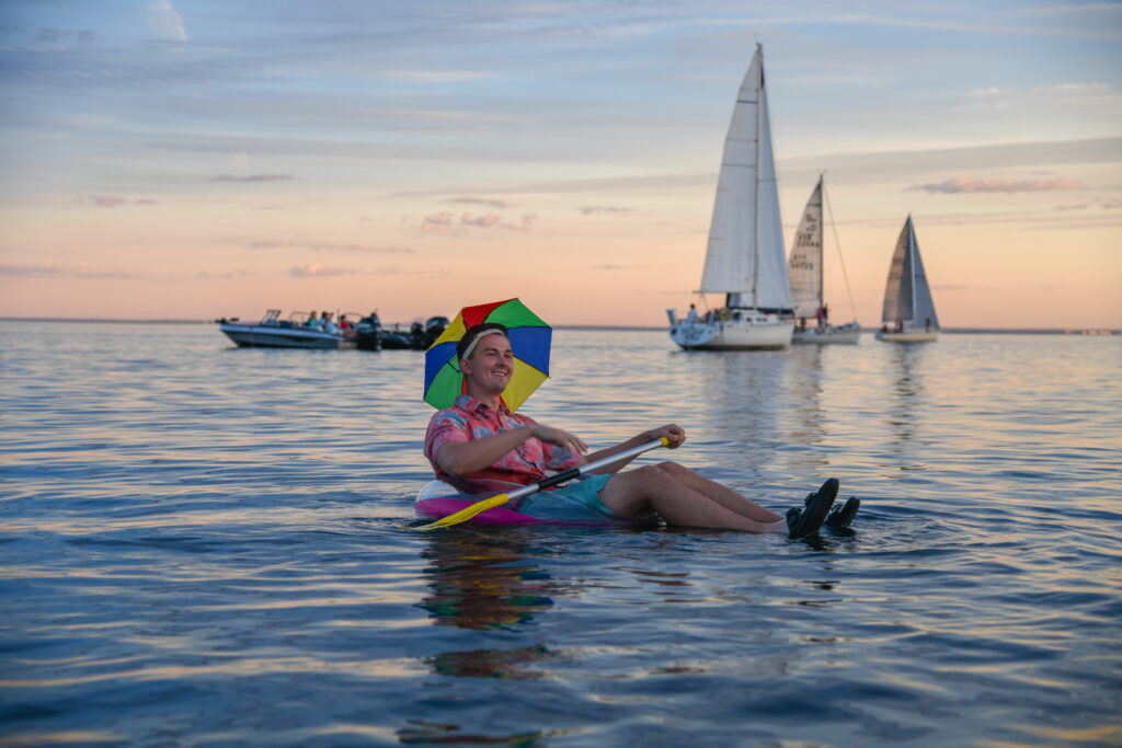 Andrew, a concertgoer, sits in the water on a floatie with a rainbow hat on and a paddle in hand. The sun is setting in the background and a few sailboats behind him.