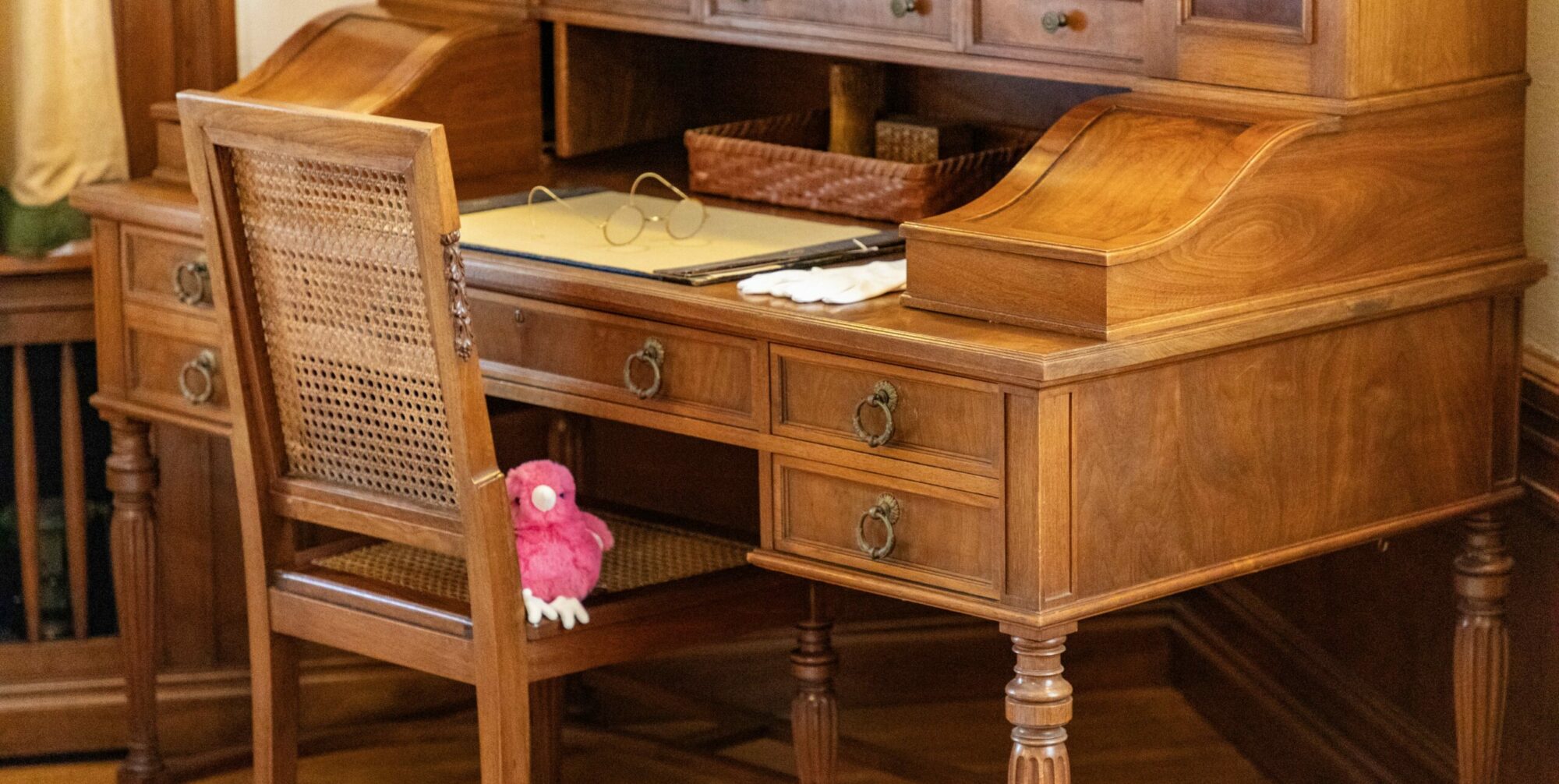 A picture of a brown wooden desk with a chair pulled up at it and a pink plush baby chick sitting on the chair.